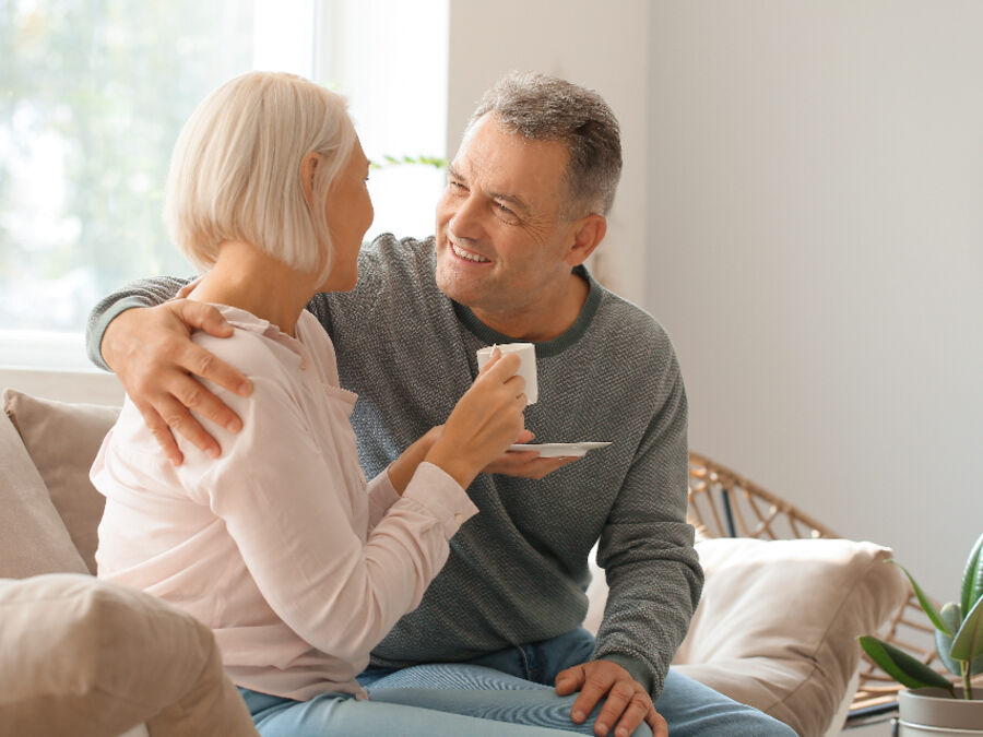 Casal de séniores sentado no sofá de casa, a conversar e a usar o telemóvel